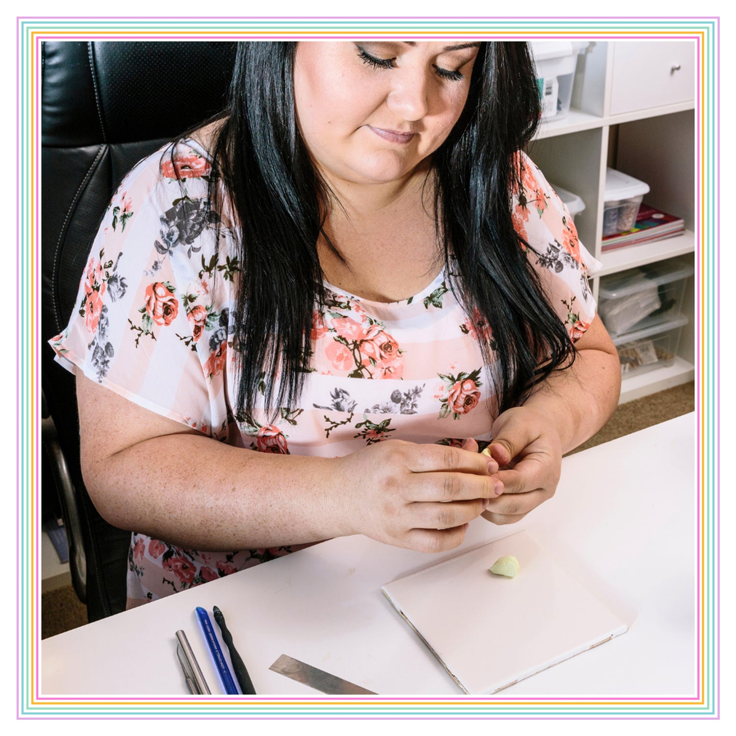 Person seated at a desk, crafting with small tools and clay. Wearing a floral top, they focus on their work. Supplies and storage visible in the background.