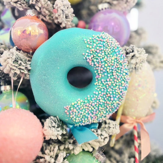 A blue frosted donut ornament with sprinkles hangs on a snowy Christmas tree, surrounded by pastel baubles.