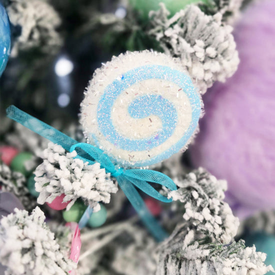 A blue and white swirl lollipop ornament with a blue ribbon is hanging on a snowy artificial Christmas tree.