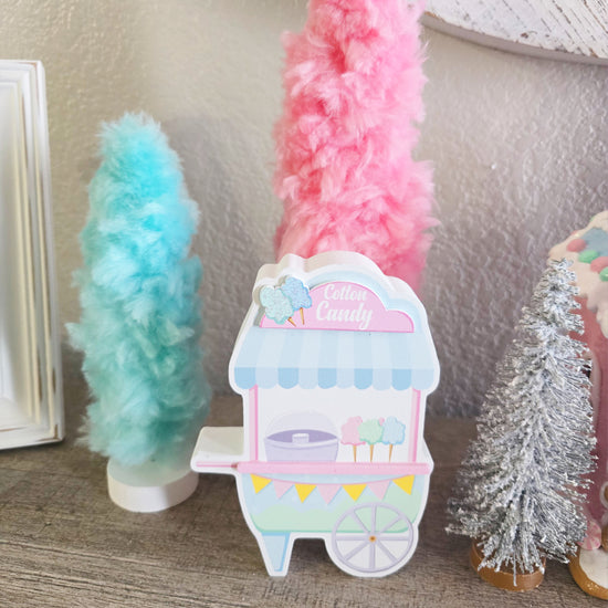 A pastel-colored cotton candy cart decoration is displayed on a wooden surface with faux cotton candy trees and a small silver tree in the background.