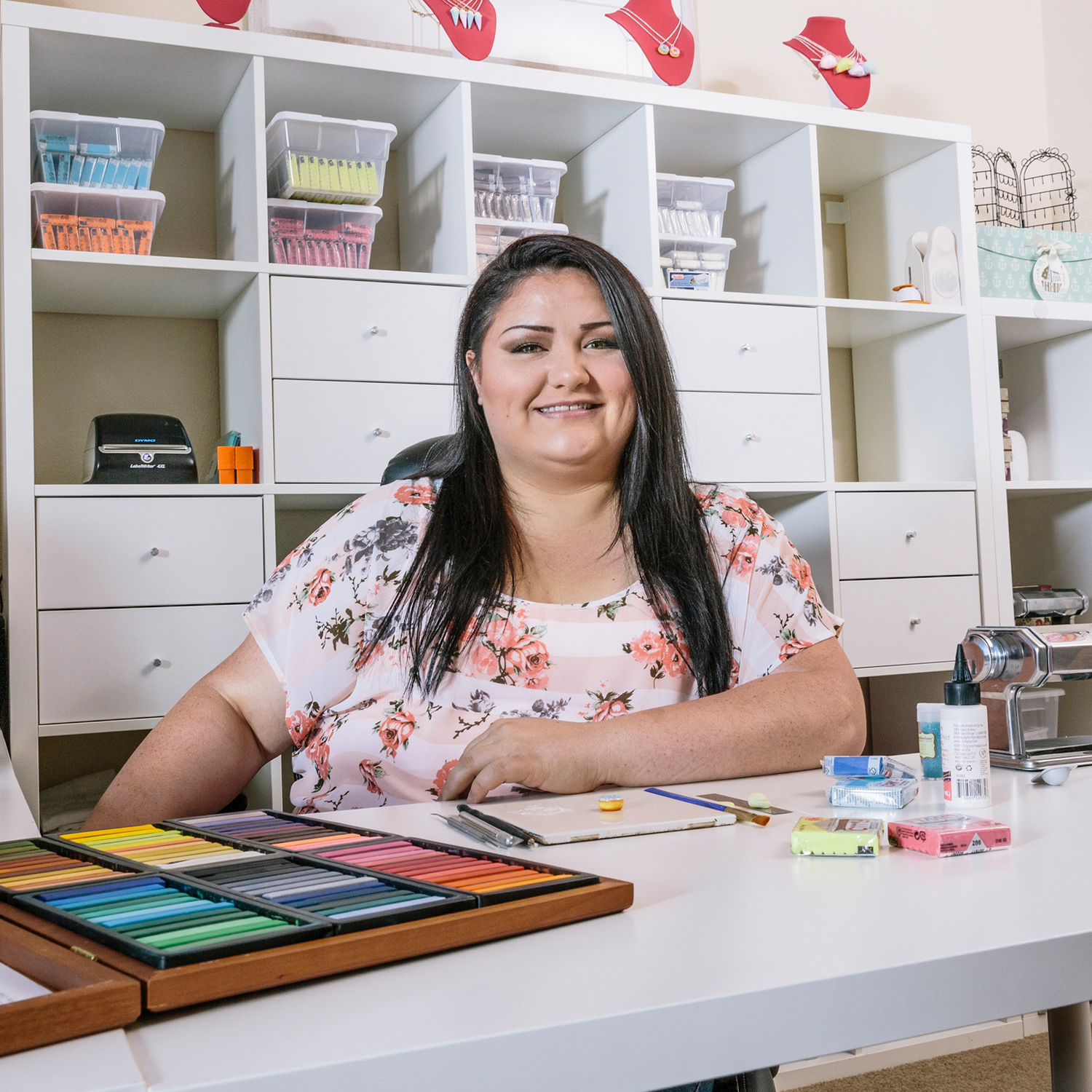 A woman sits at a desk in a craft room, surrounded by art supplies and colorful clay. Shelves with organized materials are in the background.