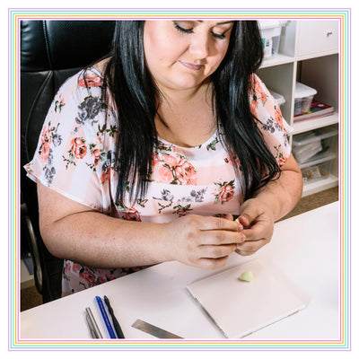 Person seated at a desk, crafting with small tools and clay. Wearing a floral top, they focus on their work. Supplies and storage visible in the background.