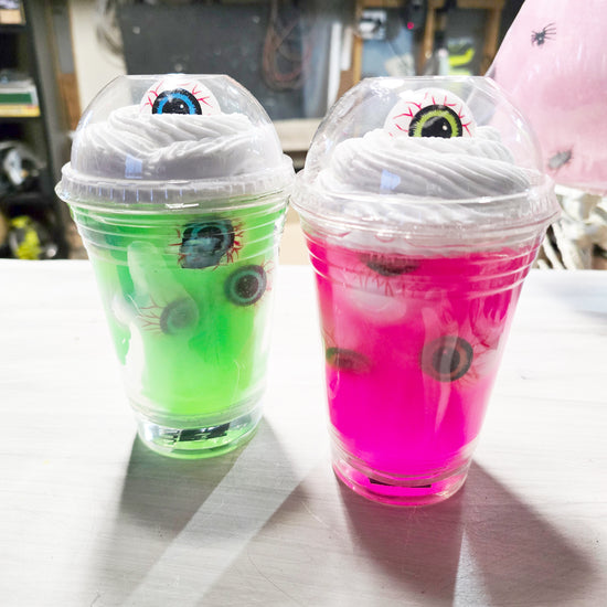 Two plastic cups with whipped cream and gummy eyeballs on top. One cup has green liquid and the other has bright pink liquid. Both have clear dome lids with eyeball stickers.