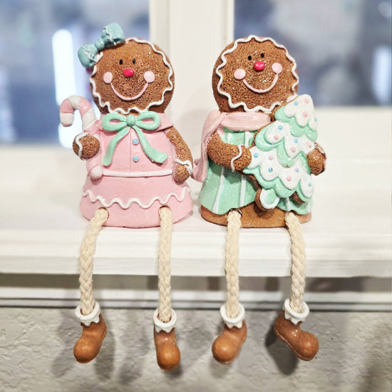 Two gingerbread figurines with smiling faces sit side by side on a ledge; one holds a candy cane, the other holds a Christmas tree. Both have rope legs and are decorated in pastel colors.