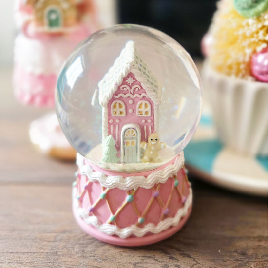 A pink snow globe featuring a small pastel house, a white tree, and a snowman, with a pink and white decorated base, sits on a wooden surface.