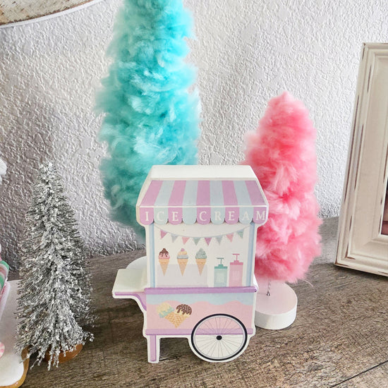 A pastel-colored miniature ice cream cart is placed on a surface, surrounded by pastel blue, pink, and silver decorative trees against a textured white wall.