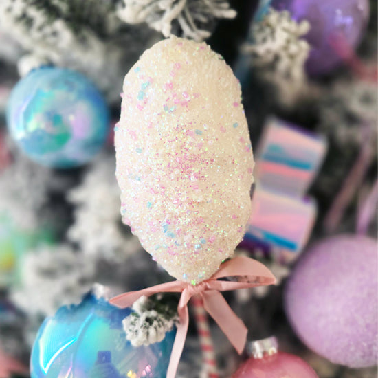 A close-up of a white, glittery ornament with pastel sparkles and a pink ribbon hanging on a decorated Christmas tree with other colorful ornaments.