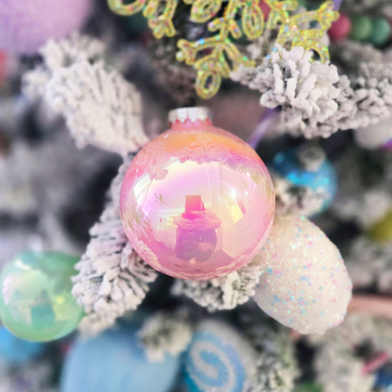A close-up of a pink reflective ornament hanging on a snow-covered Christmas tree with colorful decorations in the background.