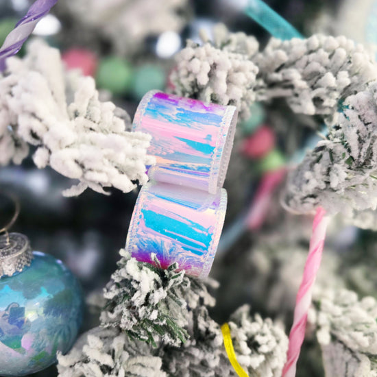Iridescent ribbon and a blue ornament hang on a snow-covered Christmas tree branch with pastel decorations.