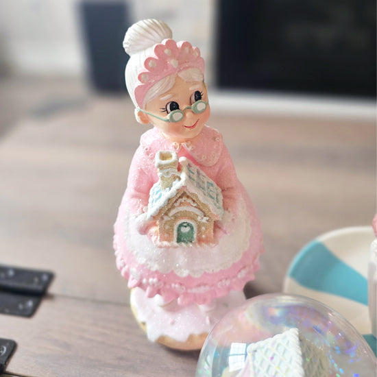 A ceramic figurine of an elderly woman with glasses and a pink dress holding a small gingerbread house stands on a wooden table.