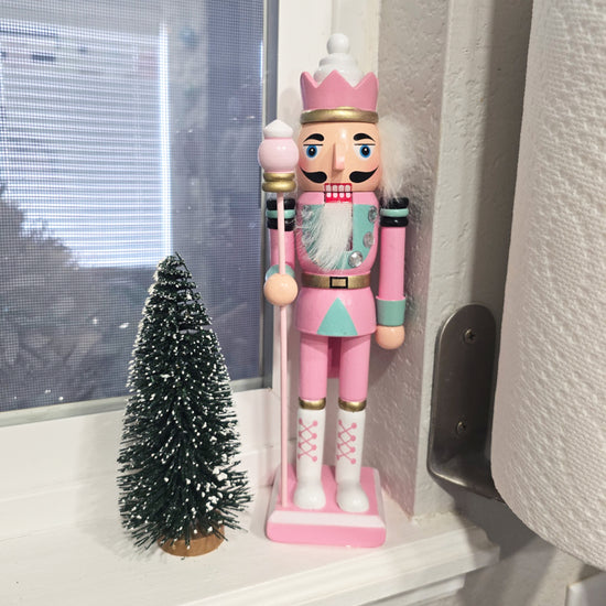 A small artificial pine tree and a pink nutcracker figurine stand on a white windowsill next to a curtain.
