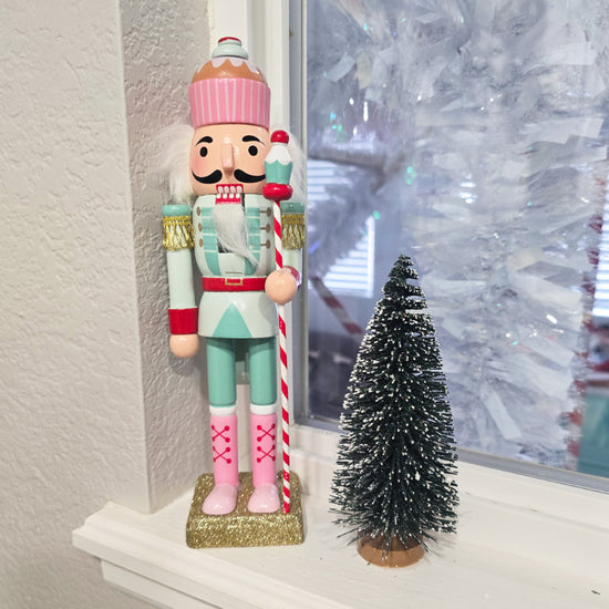 A pastel-colored nutcracker and a small artificial pine tree are displayed on a windowsill next to a frosted window.