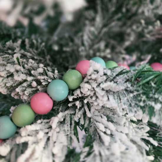 Close-up of pastel pink, green, and blue beads draped on snow-dusted artificial pine branches.
