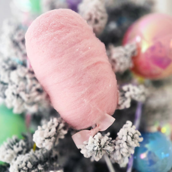 A close-up of pink cotton candy on a frosted, snow-dusted branch with colorful, blurred ornaments in the background.