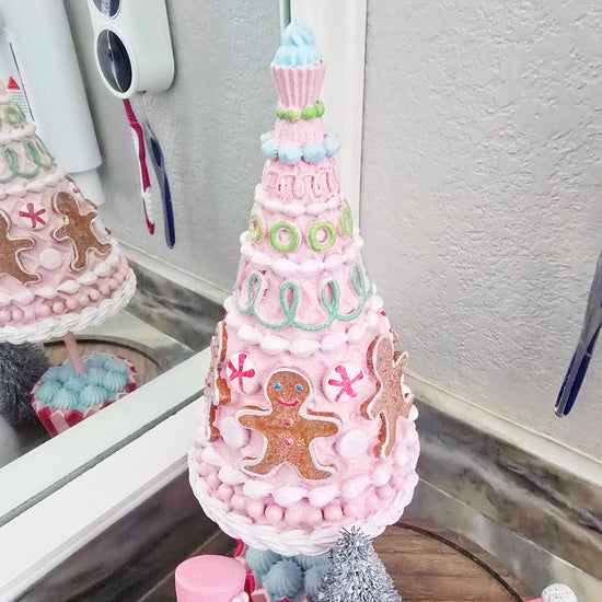 A pink, cone-shaped decorative tree with gingerbread and candy designs sits on a bathroom counter near a mirror.