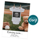 A baby onesie with an ultrasound image hangs on a clothesline at a baseball field, with a teddy bear, baseball, and glove nearby. Text: "Coming soon... Baby Hall due January 2026.