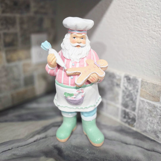 A figurine of Santa Claus dressed as a baker holding a piping bag and a gingerbread cookie, standing on a marble countertop near a tiled wall.