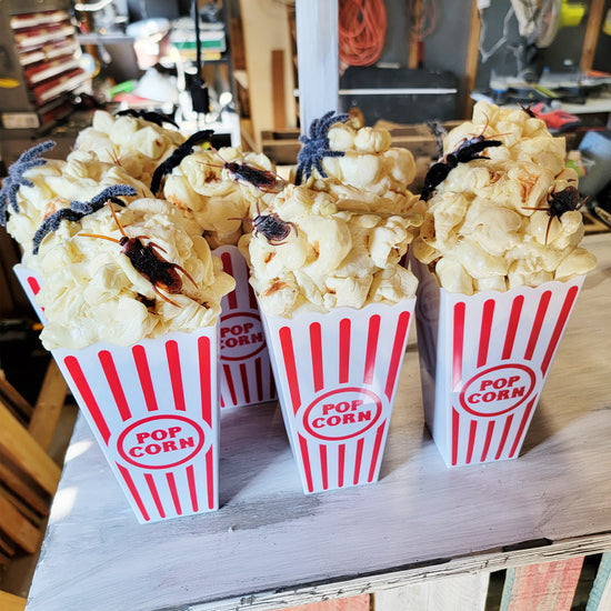 Popcorn boxes filled with popcorn and decorated with toy spiders and cockroaches on a wooden surface.