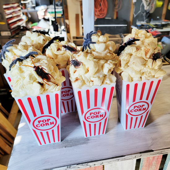 Popcorn containers filled with fake popcorn and topped with fake black spiders and cockroaches, placed on a wooden surface.