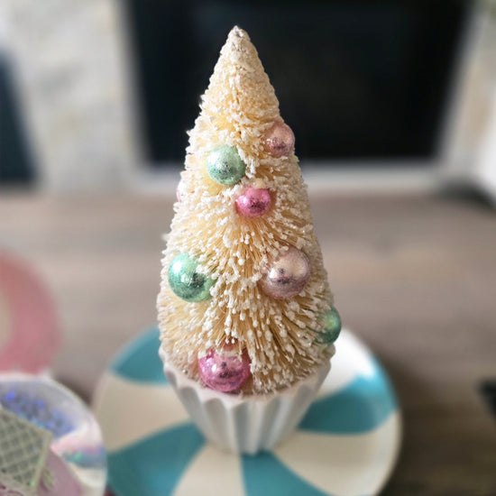 A small, white bottle brush Christmas tree decorated with pastel ornaments sits in a cupcake-shaped holder on a striped plate.