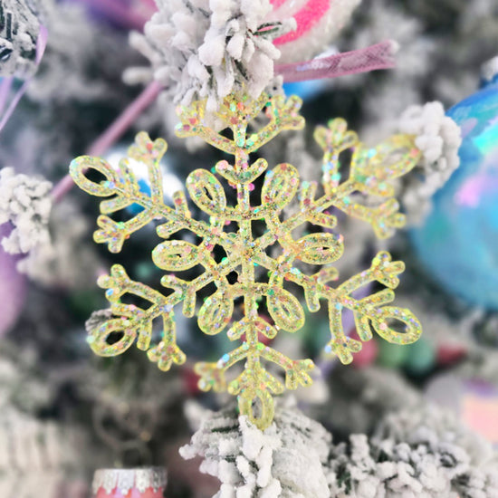 A glittery yellow snowflake ornament hangs on a snow-covered Christmas tree with other colorful decorations in the background.